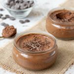 Deux ramequins de mousse au chocolat, décorées de petites pépites de chocolat, posées près d'une cuillère.