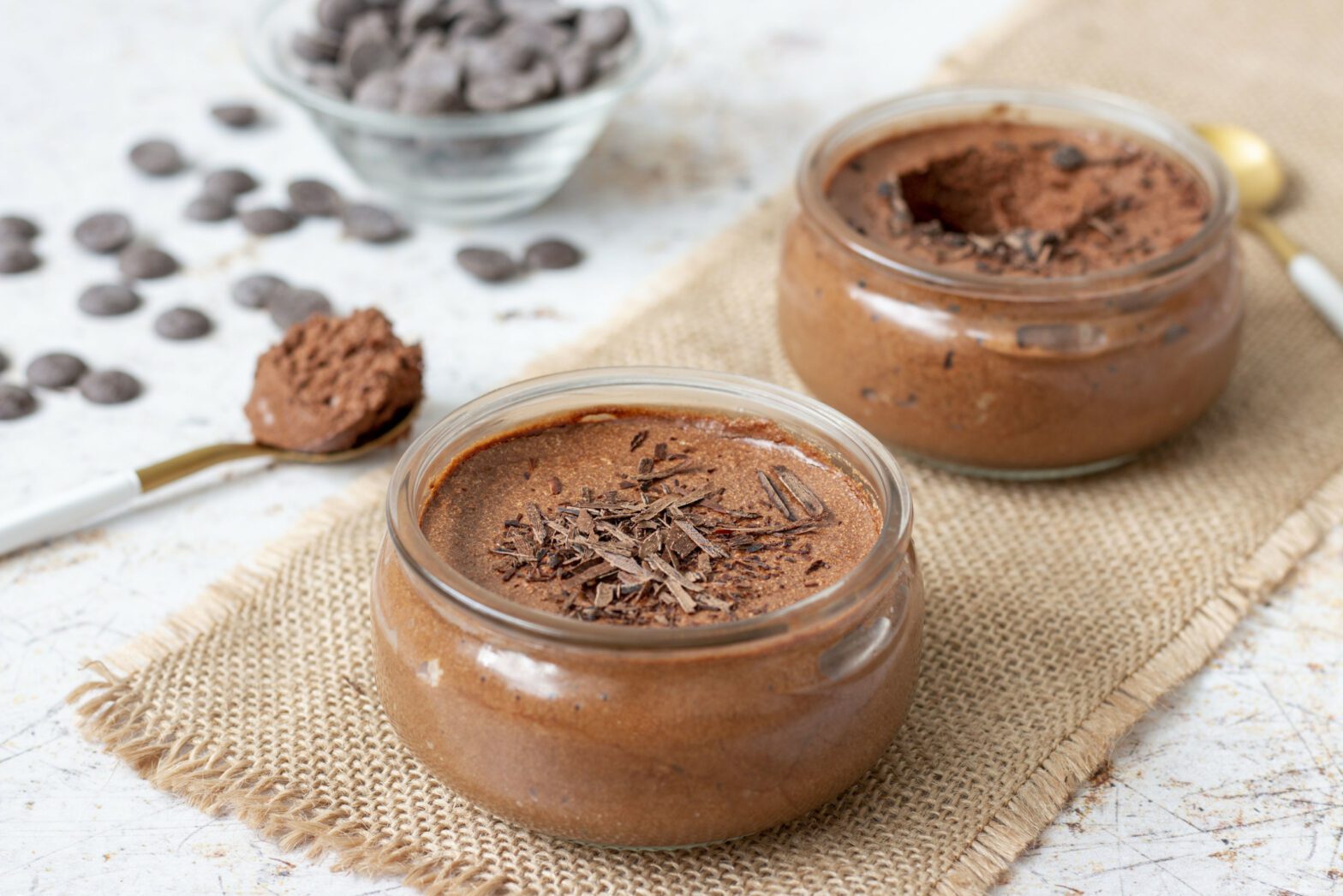 Deux ramequins de mousse au chocolat, décorées de petites pépites de chocolat, posées près d'une cuillère.