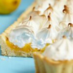 Part de tarte garnie de crème de citron et de meringue dorée, entourée de citrons disposés sur une table bleue